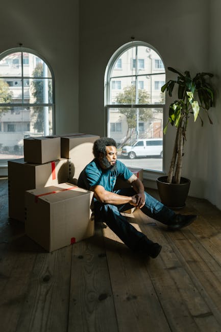 A man with a curly black afro and a beard is seen packing items into a cardboard box labeled 'Caution This Side Up' in a well-lit indoor space, likely preparing for a home relocation or furniture tran