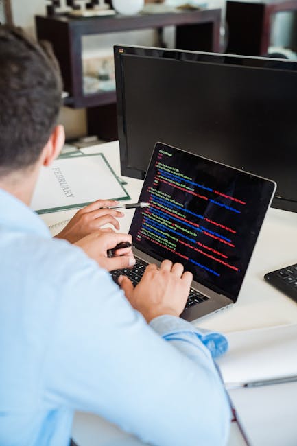A person seated at a desk inside a home or office environment, engaged in a packing or moving preparation process. The individual is wearing a light blue shirt and is using a laptop displaying lines o