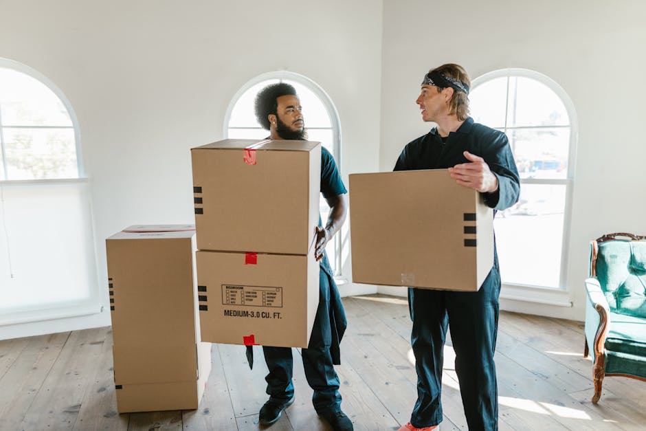 A person seated at a desk inside a home or office environment, engaged in a packing or moving preparation process. The individual is wearing a light blue shirt and is using a laptop displaying lines o