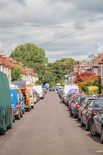 A residential street lined with parked cars on both sides, including a variety of sedans, hatchbacks, and vans. Some vehicles are partially obscured by building structures, while others are clearly vi